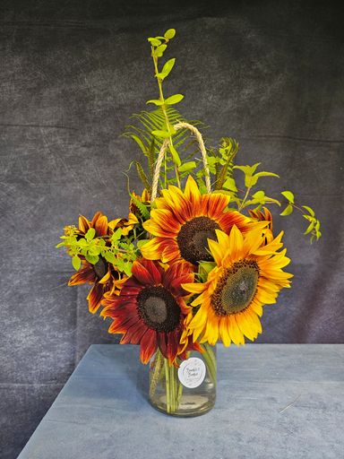 Bright yellow, orange and red short sunflowers with green foliage, arranged in a jar with a round sticker on the front and jute rope handle.