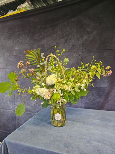 A glass jar with a round sticker and jute rope handle containing white and pink/peach-coloured fresh-cut flowers and hedgerow foliage.  