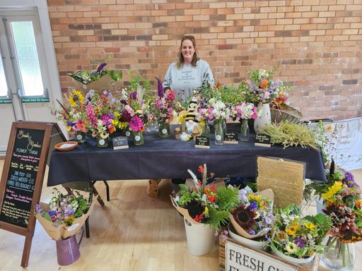 A lady behind a table with a black tablecloth. On the table are jars of fresh flowers, and bouquets in buckets are in front of the table. 
