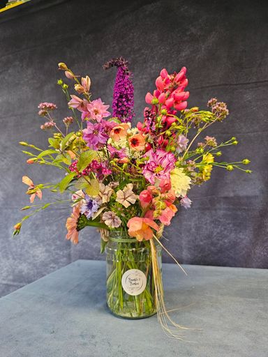 A large jar with a round white sticker on the front containing bright pink, orange, yellow, and purple tall flowers with green foliage.  