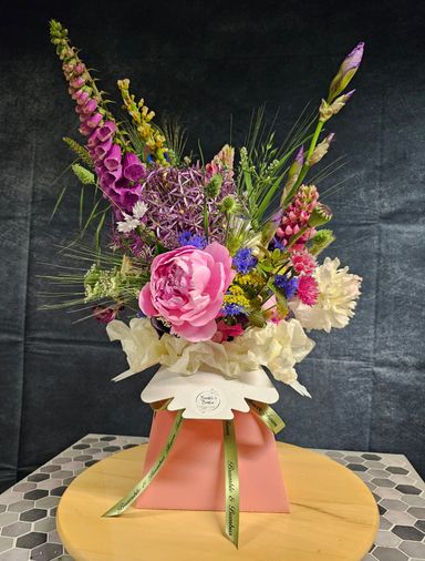 Tall pink, purple and white flowers with green foliage in a pink card Living vase with a green ribbon tied. In front of a black background. 
