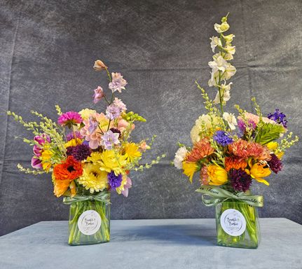 Two short square jars side by side with a white round sticker on the front.   Full of bright yellow, red, purple and orange flowers.  